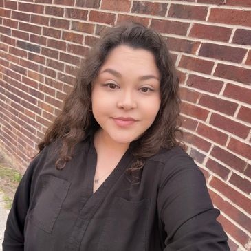 A woman with curly hair wearing a black top stands outdoors by a brick wall.