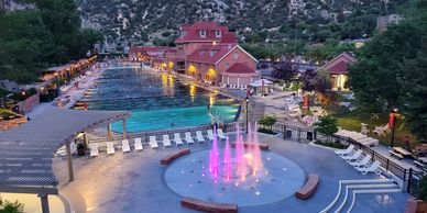 Evening view of a lit water fountain and pool area by red buildings in a mountainous setting.