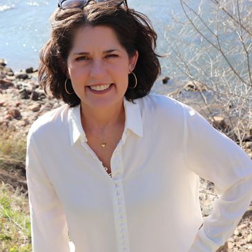 Smiling woman in white blouse outdoors near rocky water edge.