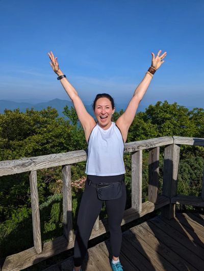 Woman joyfully posing with arms raised on a wooden deck with scenic greenery.