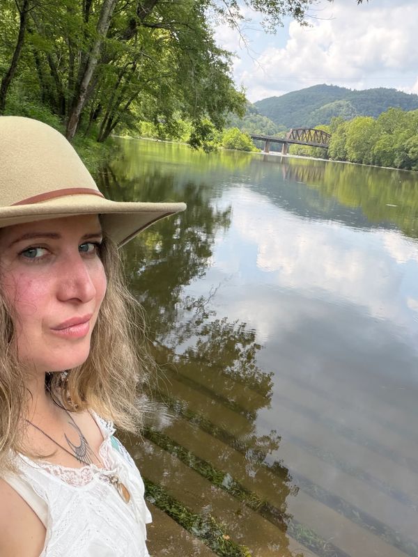 Woman in a hat by a river with a bridge in the background.