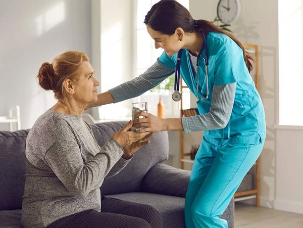 Nurse giving water to elderly woman sitting on couch.