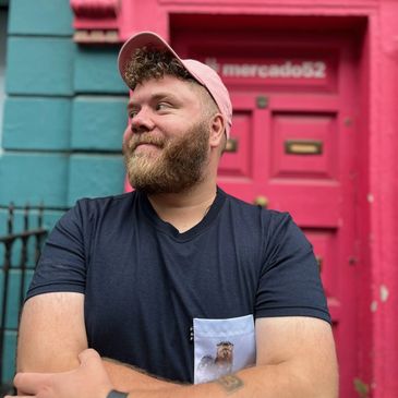 Man with a beard and pink cap stands smiling against a vibrant pink door.