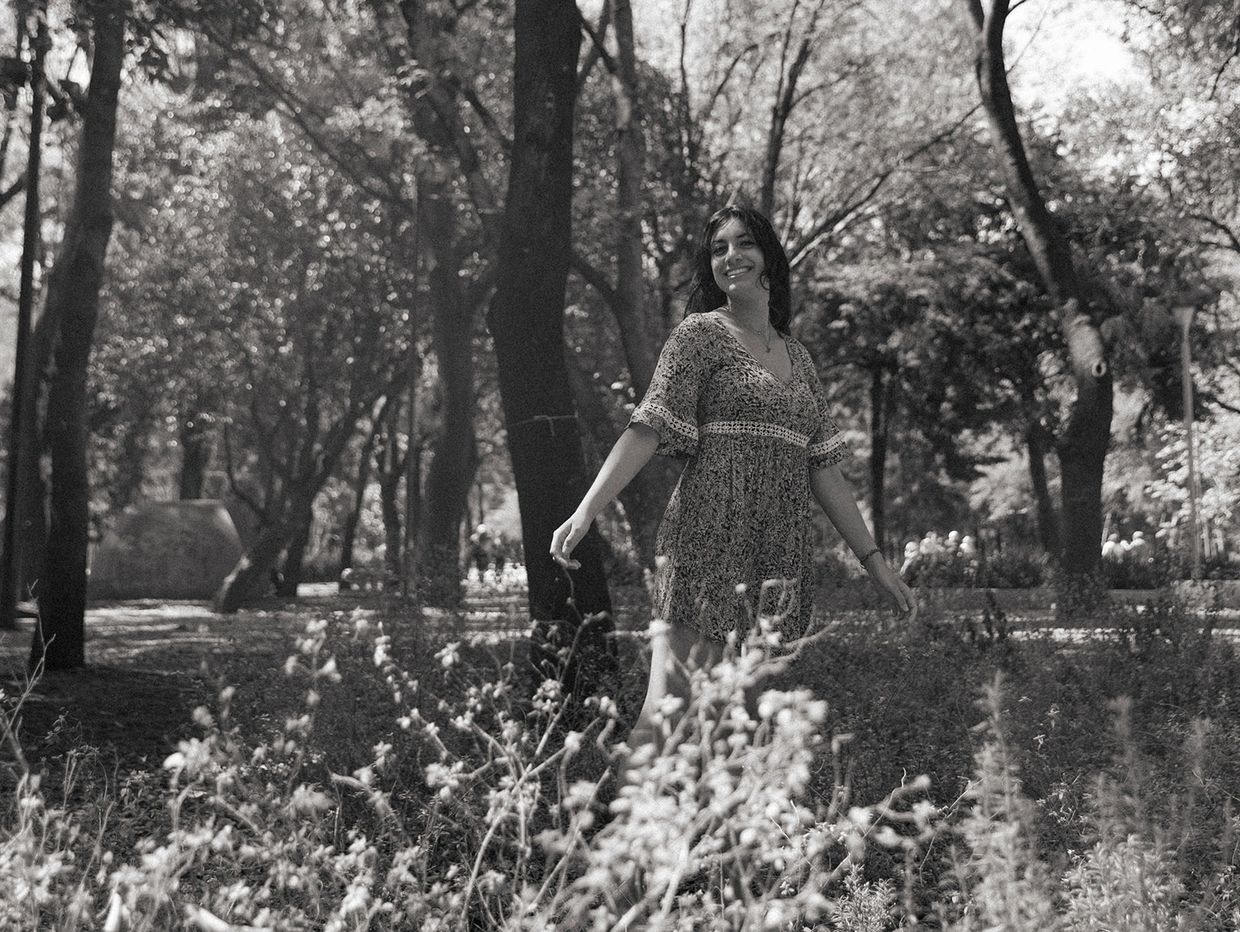 A woman in a floral dress smiles while standing in a sunlit forest clearing.