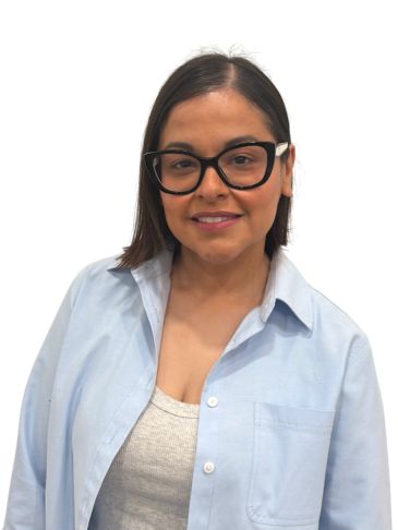 A woman with glasses wearing a light blue shirt and a white top smiles.