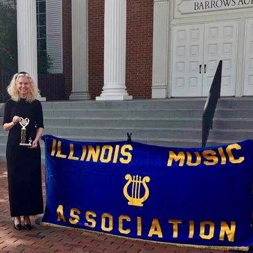 Woman in black dress holding trophy beside Illinois Music Association banner.