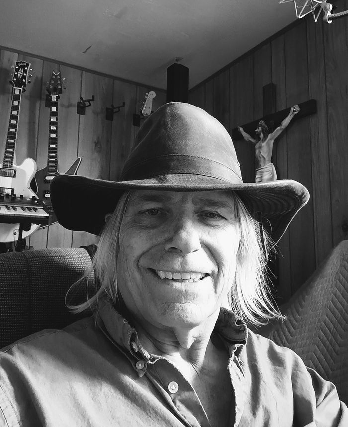 Smiling man wearing a wide-brimmed hat in a wood-paneled room with guitars and a crucifix.