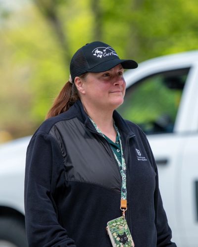 Woman in casual outdoor clothing and cap standing near a white vehicle.