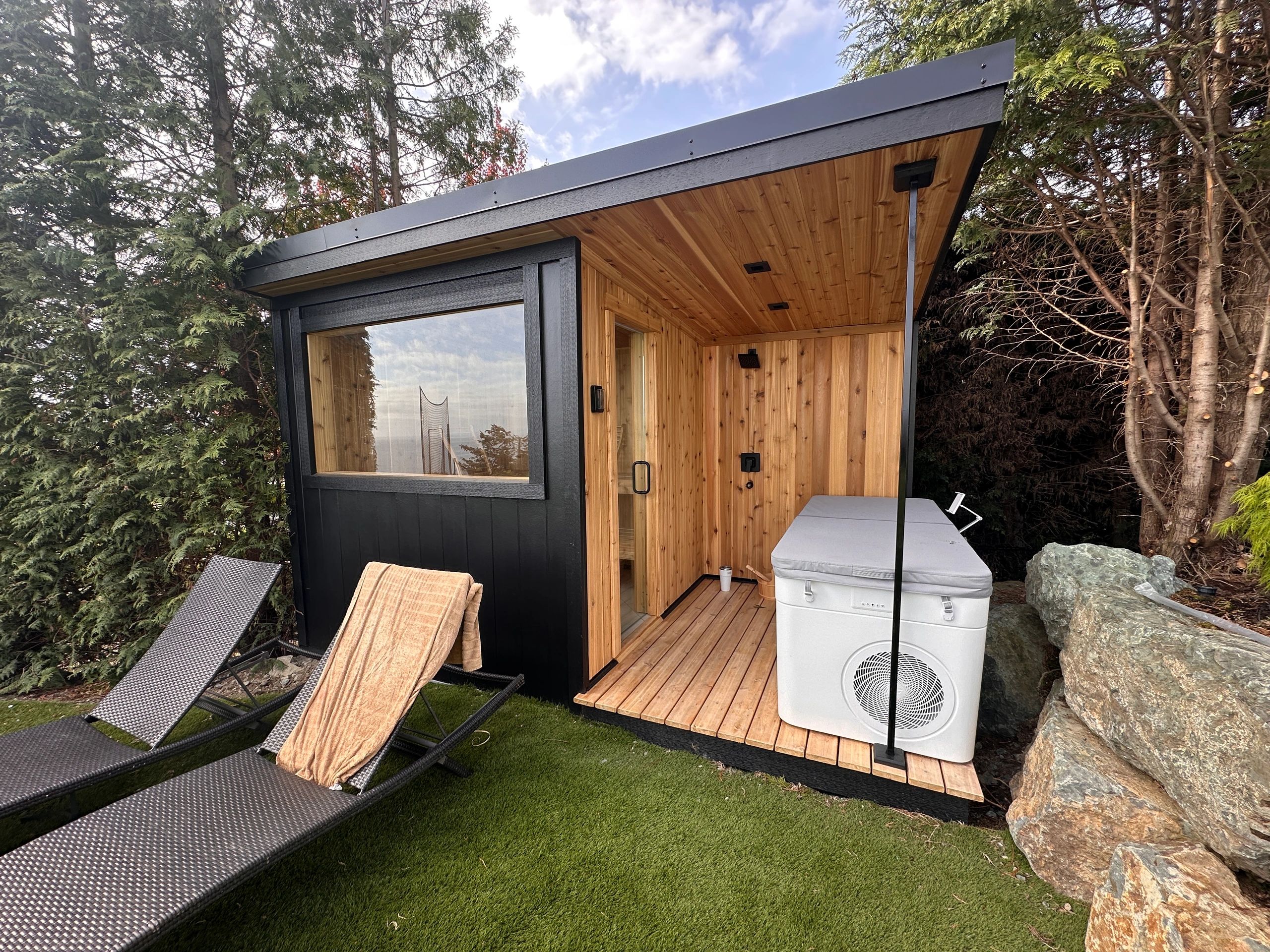 Modern outdoor wooden sauna with hot tub and lounge chairs surrounded by trees.