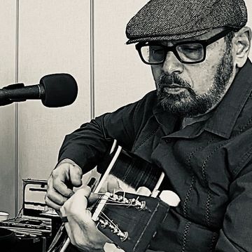 Man with glasses and cap playing guitar near microphone.
