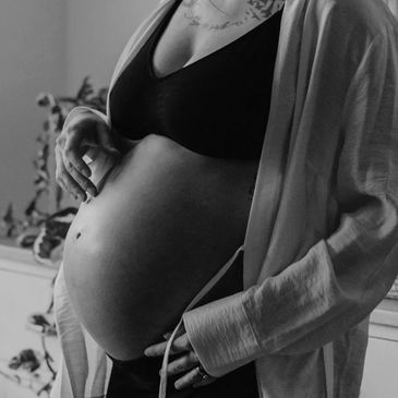 Pregnant woman gently touching her belly, wearing a black top and light robe indoors.