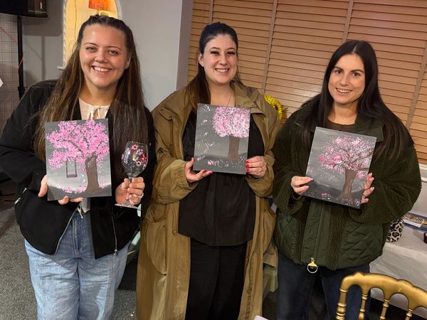 Three women proudly display their cherry blossom paintings and decorated wine glasses indoors.