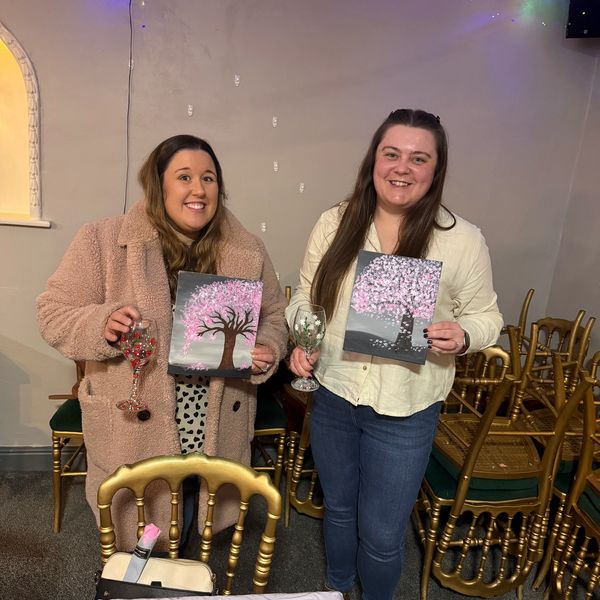 Two women proudly display their cherry blossom tree paintings with decorated wine glasses.
