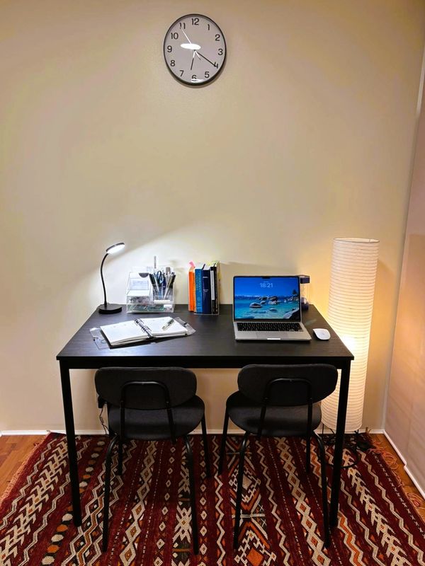 A tidy study desk with two chairs, a laptop, books, lamp, and clock on a patterned rug.