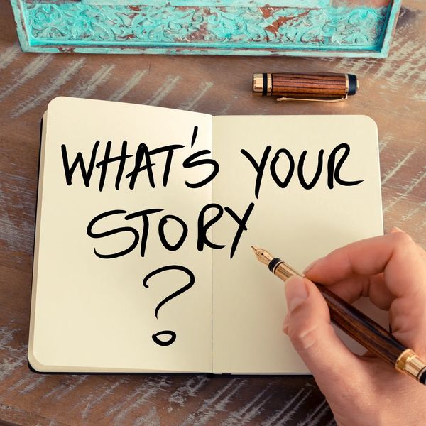 An open journal with What's Your Story written in black ink with a hand holding a pen