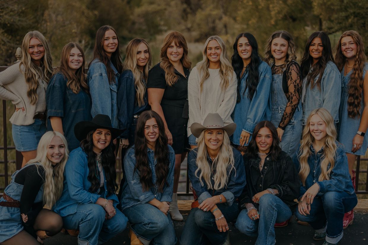 Group of smiling women, many in denim, posing outdoors with a nature backdrop.