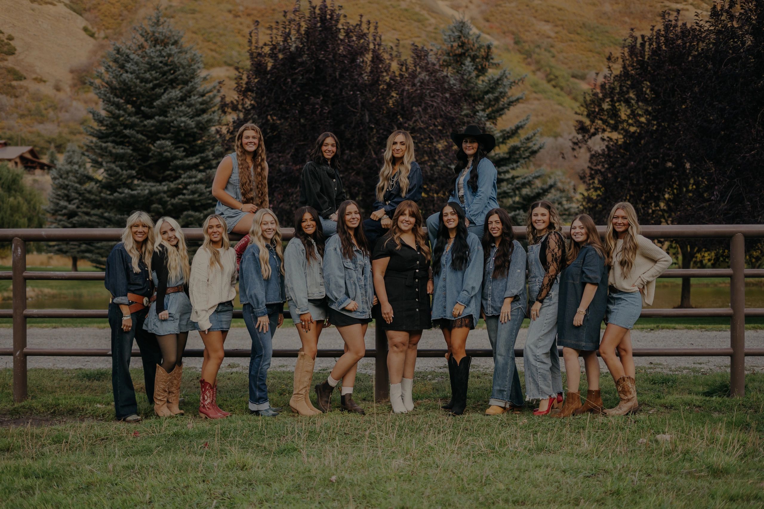 A group of women posing outdoors with trees and hills in the background.