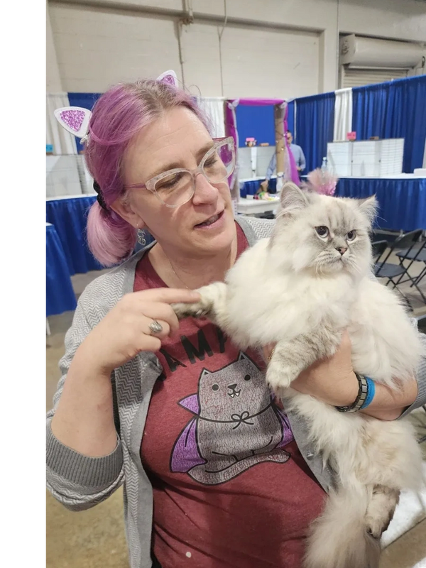 Woman with purple hair holds a fluffy cat at an indoor event.