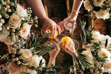 Four hands holding glasses of orange juice surrounded by white rose bouquets.