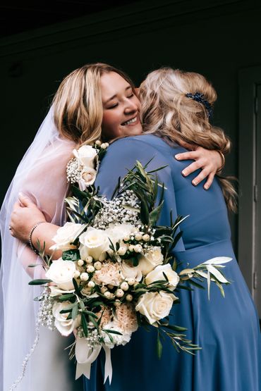 Bride and woman in blue dress hugging warmly with bouquet of white flowers.