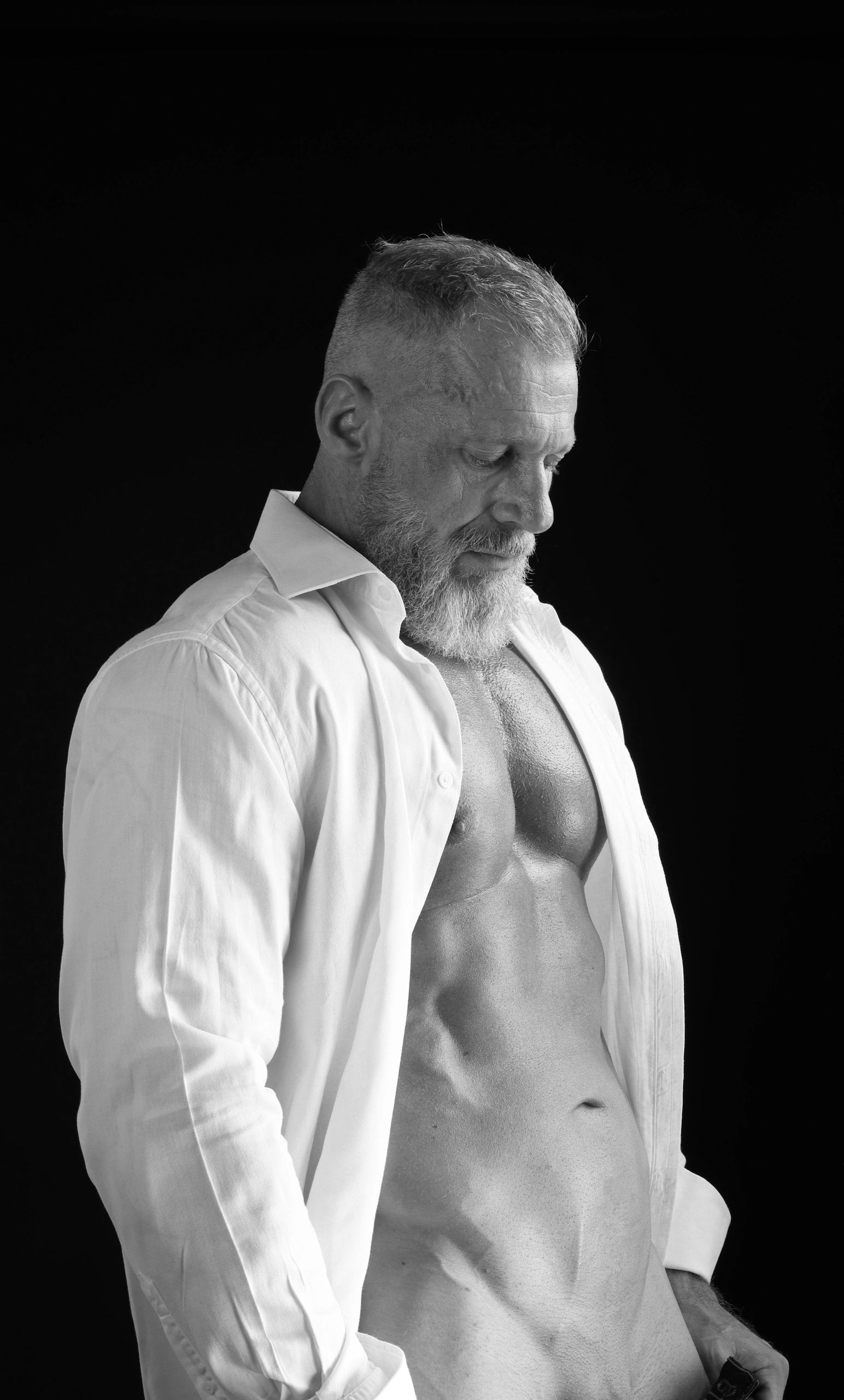 A muscly man, with a white cotton shirt, bare tanned chest. The shirt is open. He is looking down. 