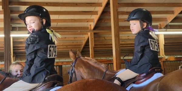 Two young riders in helmets and jackets on horses inside a stable.