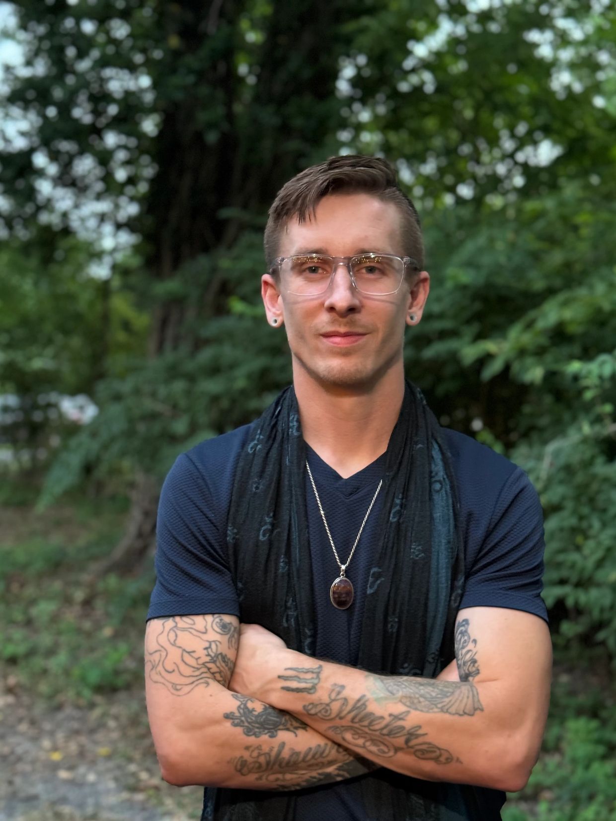 Man with tattoos and glasses stands confidently in nature with arms crossed.