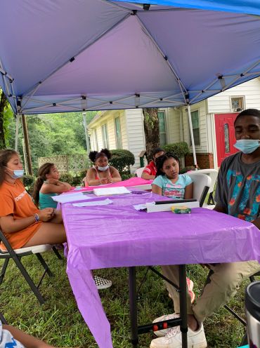 Kids and a young man seated under a canopy with a purple tablecloth.