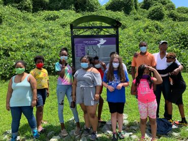Group wearing masks outdoors near a green hill.