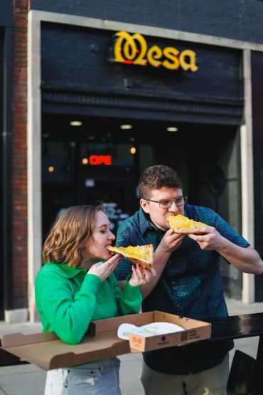 Two people enjoying pizza outside a restaurant named Mesa.