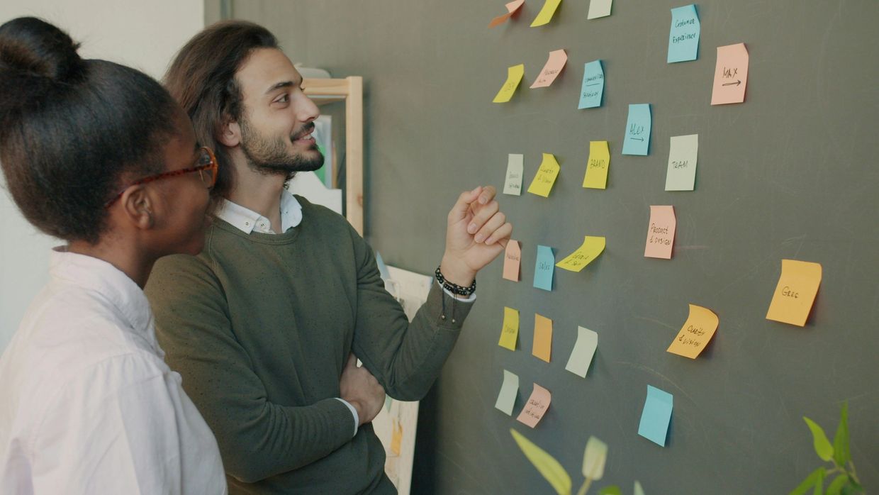 Two colleagues discuss ideas using colorful sticky notes on a blackboard.