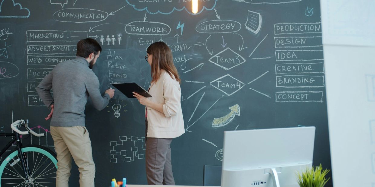 Two coworkers brainstorming and writing on a large chalkboard wall filled with marketing and strategy notes.