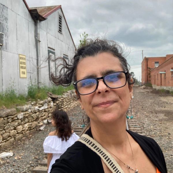 Woman with glasses smiling near railroad tracks and buildings on a cloudy day.
