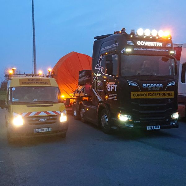 Two large trucks with "Convoi Exceptionnel" signs transporting oversized cargo at dusk.
