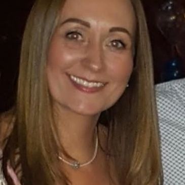 Smiling woman with straight brown hair in a dimly lit setting.