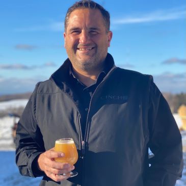 Man in black jacket holding a glass of beer outdoors on a sunny day.