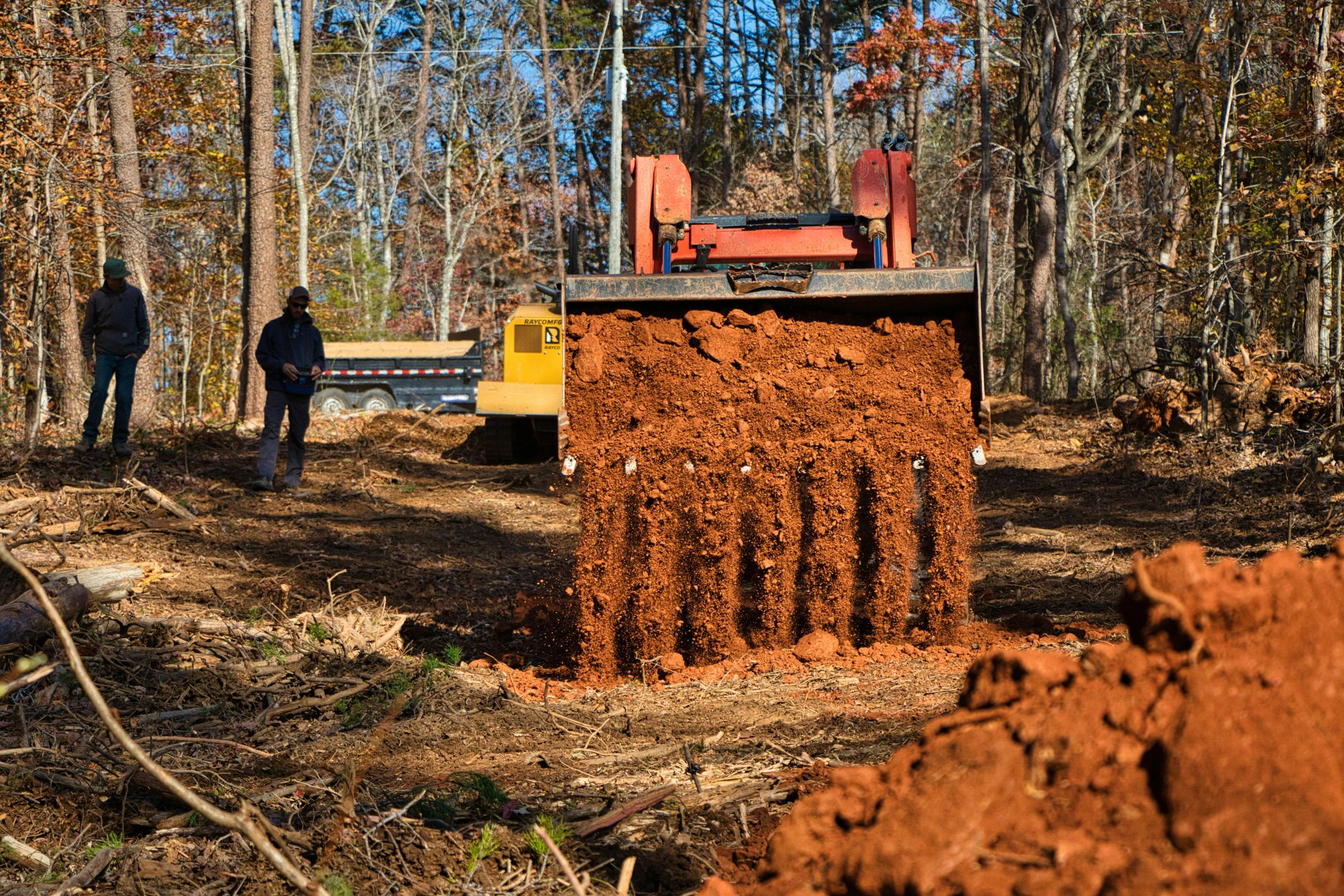 A bulldozer dumping red dirt in a forest clearing with two workers nearby.