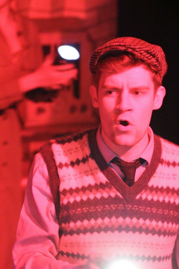 A young man in flat cap and woollen jumper looks concerned surrounded by red light
