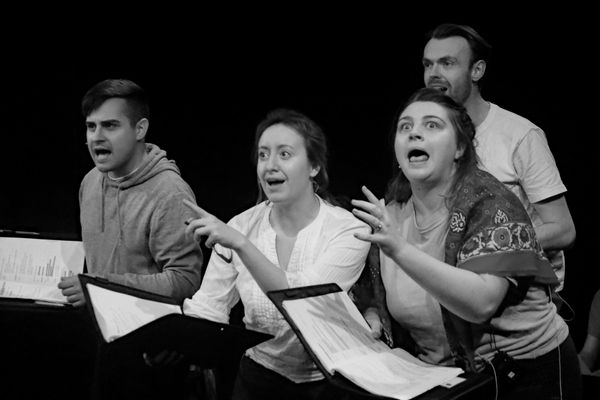 4 actors rehearse a comical scene, holding scripts and presenting exaggerated facial expressions