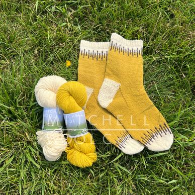 yellow hand knit socks sit on a grassy field next to a yellow & white skeins of yarn