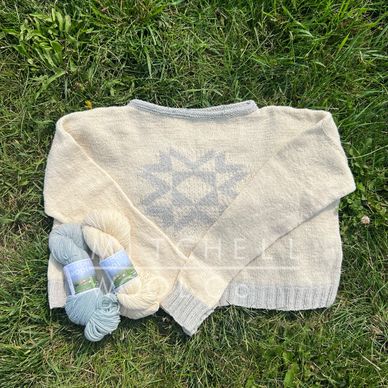 a white and bloue sweater with a quilt block motif sits on green grass