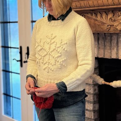 a white woman knits in front of a fireplace wearing a white sweater with a snowflake motif