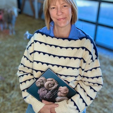 sherry holds a knitting book while wearing a blue and white sweater in the barn