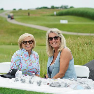 Two women smiling outdoors at a table with items.