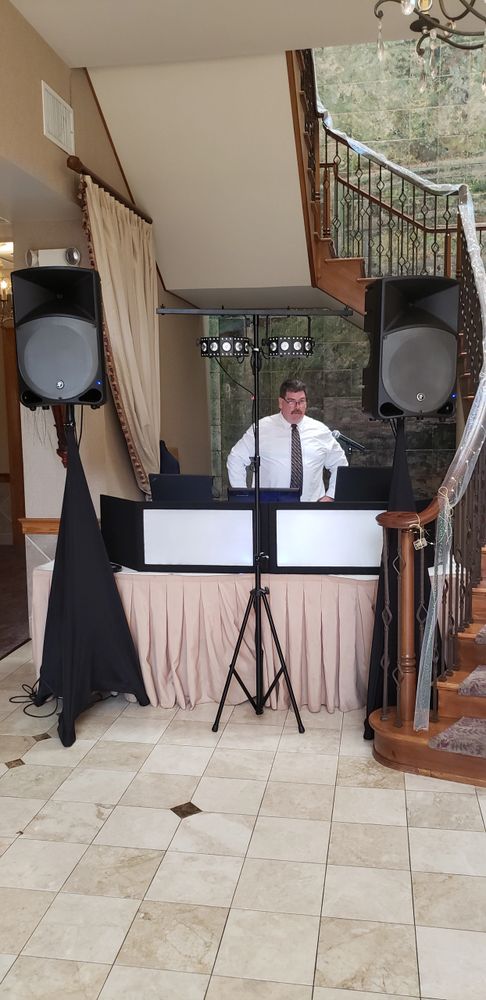 Man in white shirt standing behind DJ setup with speakers and lighting.