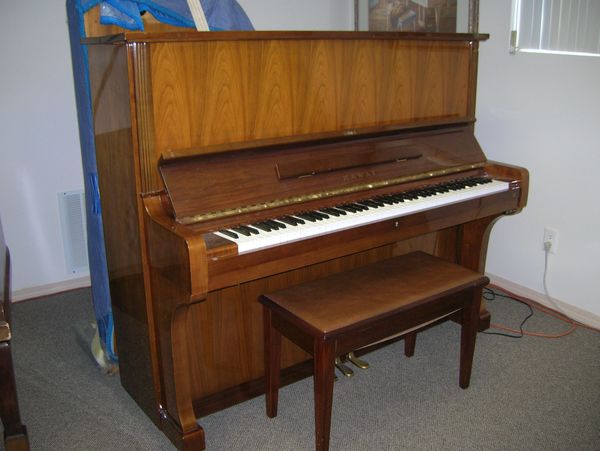 Modern walnut upright piano in storage in front of a grand piano packed up for moving.