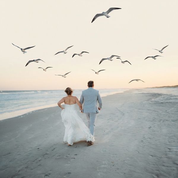 Couple in wedding attire walking on the beach with seagulls flying above.