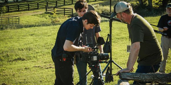 Three men adjusting camera equipment outdoors in a grassy field.