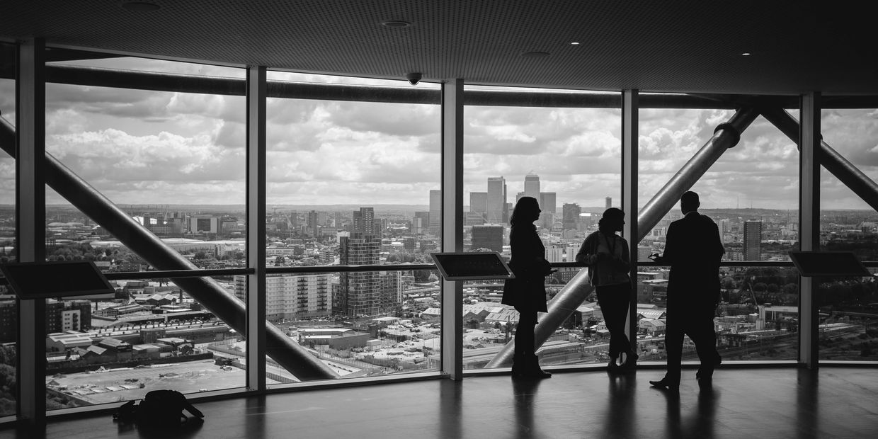 Silhouettes of three people talking by a large window overlooking a cityscape in black and white.