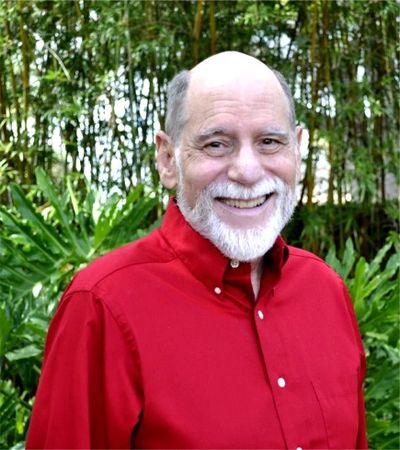Elderly man with a white beard smiling, wearing a red shirt outdoors.